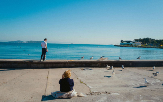 Une femme regarde l’horizon en bord de mer, avec une mouette en vol et un homme assis sur un muret de plage dans une lumière douce